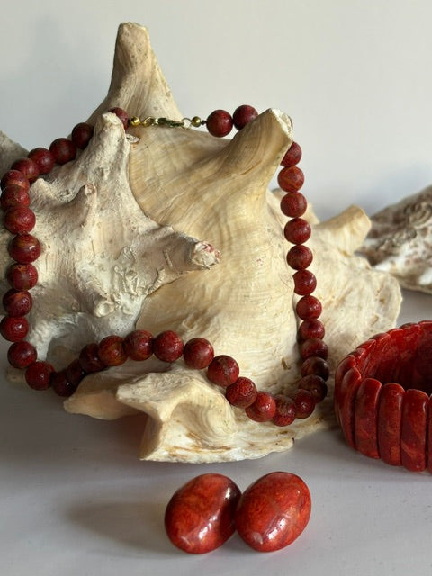 Rote Schaumkorallenkette vor einer großen Muschel mit passendem Armband und ovalen Ohrsteckern